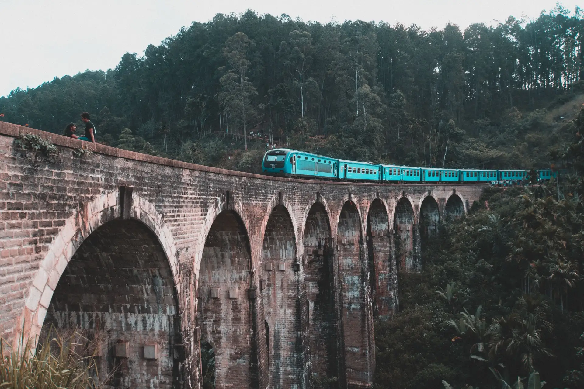 A turquoise Sri Lanka Railways train crossing the stone Nine Arch Bridge at Demodara, seen from below the arches, with two people sitting on the parapet and forested hillside behind.
