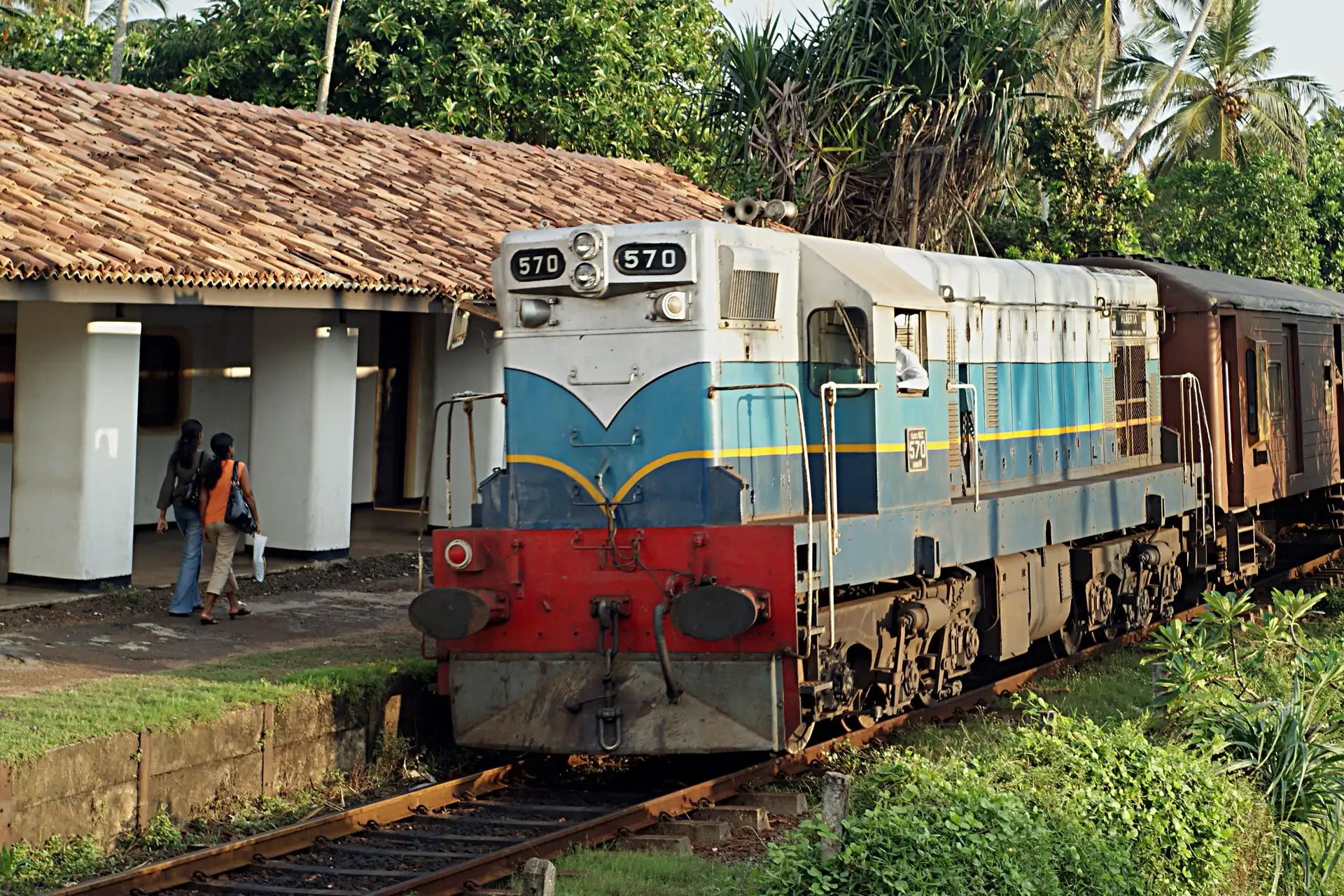 A blue, white and red diesel locomotive numbered 570 standing at the small tiled-roof platform of Bentota station under coconut palms, with two women walking along the platform.