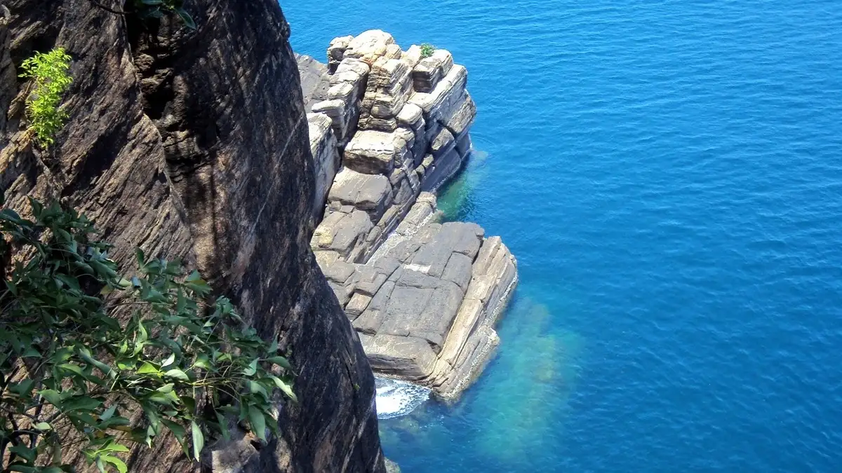 Looking straight down the dark granite cliff of Swami Rock at Trincomalee to a stack of pale layered rock and clear blue sea below.