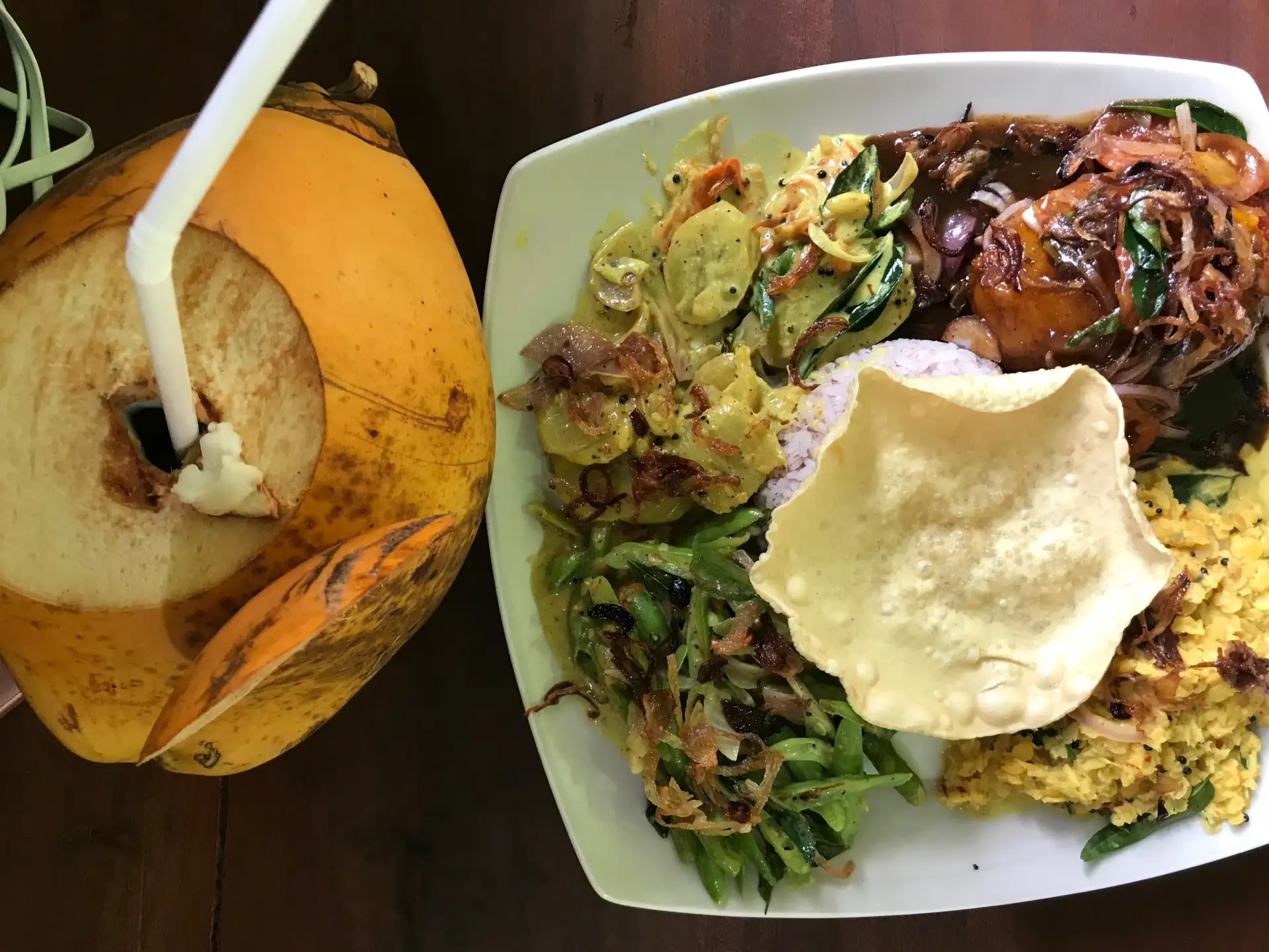 A white plate of rice and curry seen from above — dhal, mallung, aubergine moju, green beans and a papadam — beside an orange king coconut with a straw.