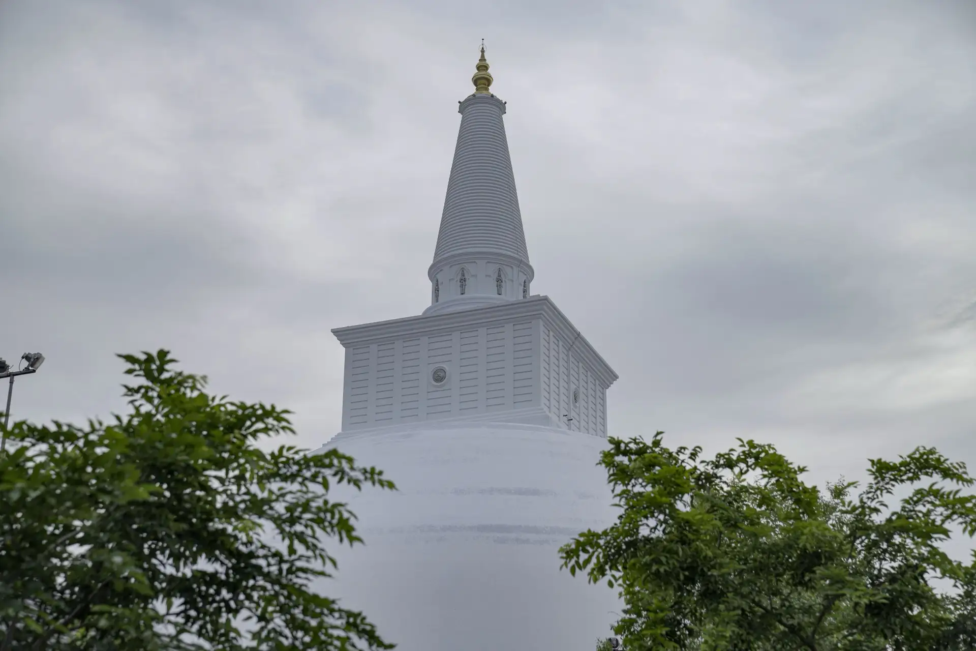 The whitewashed dome and gold spire of the Ruwanwelisaya stupa in Anuradhapura, rising above trees under grey cloud.