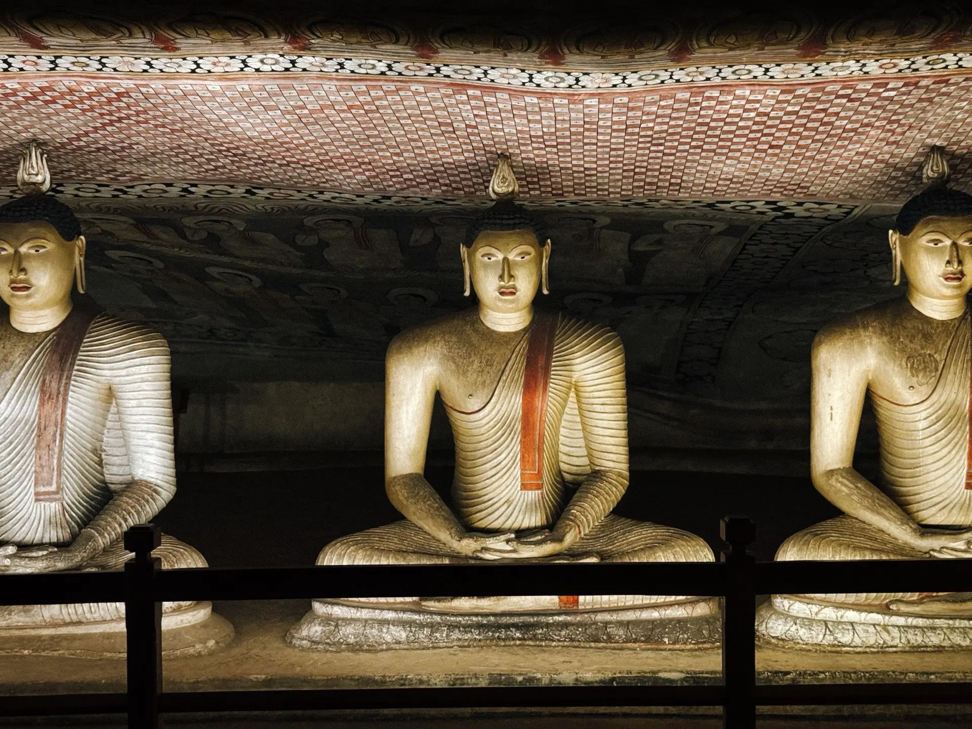 Three seated Buddha statues in a Dambulla cave, beneath a painted rock ceiling that follows the curve of the overhang.