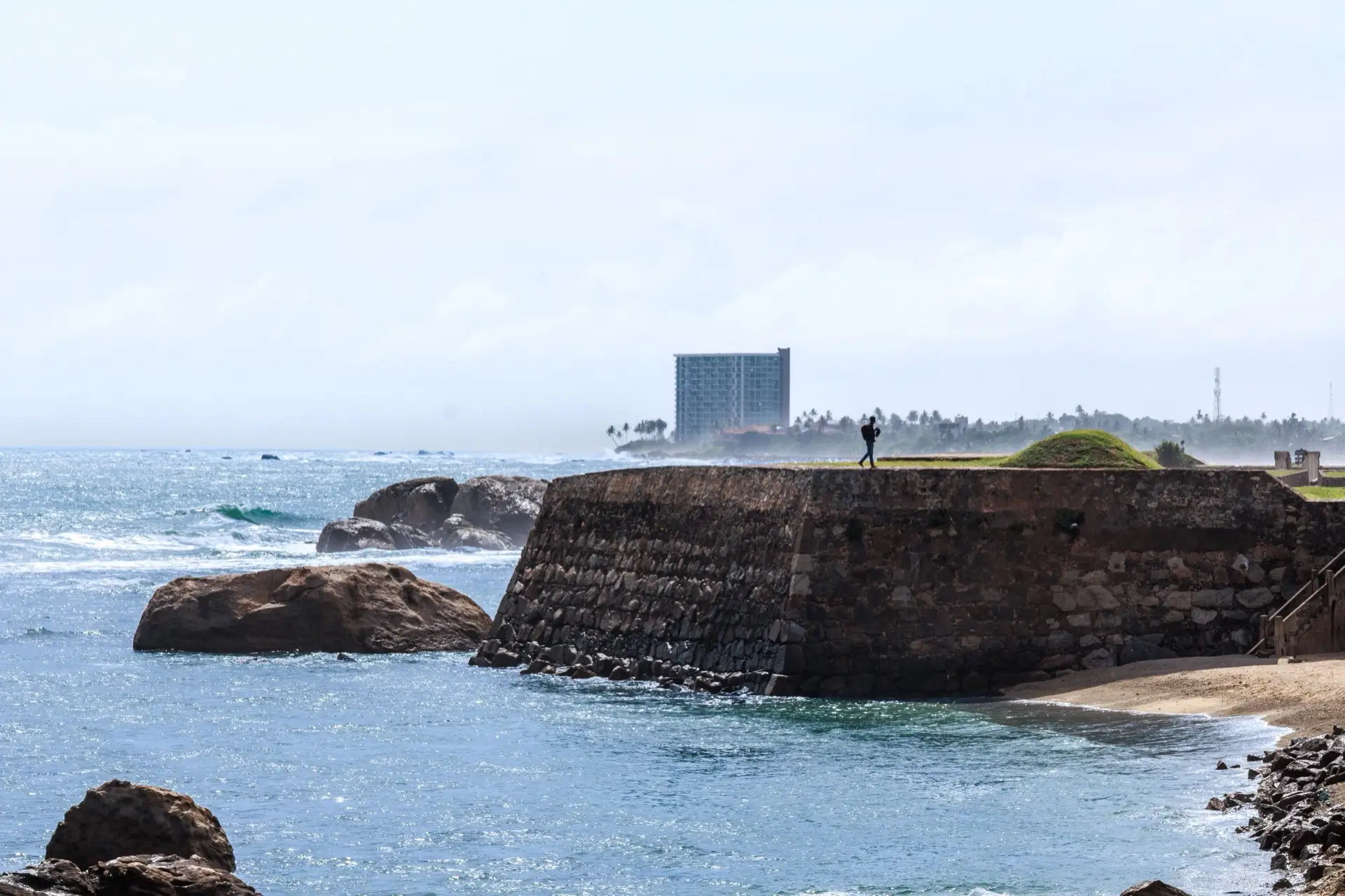 A stone bastion of the Galle Fort ramparts dropping to the sea, with one person walking the grass along the top and surf breaking on the rocks below.