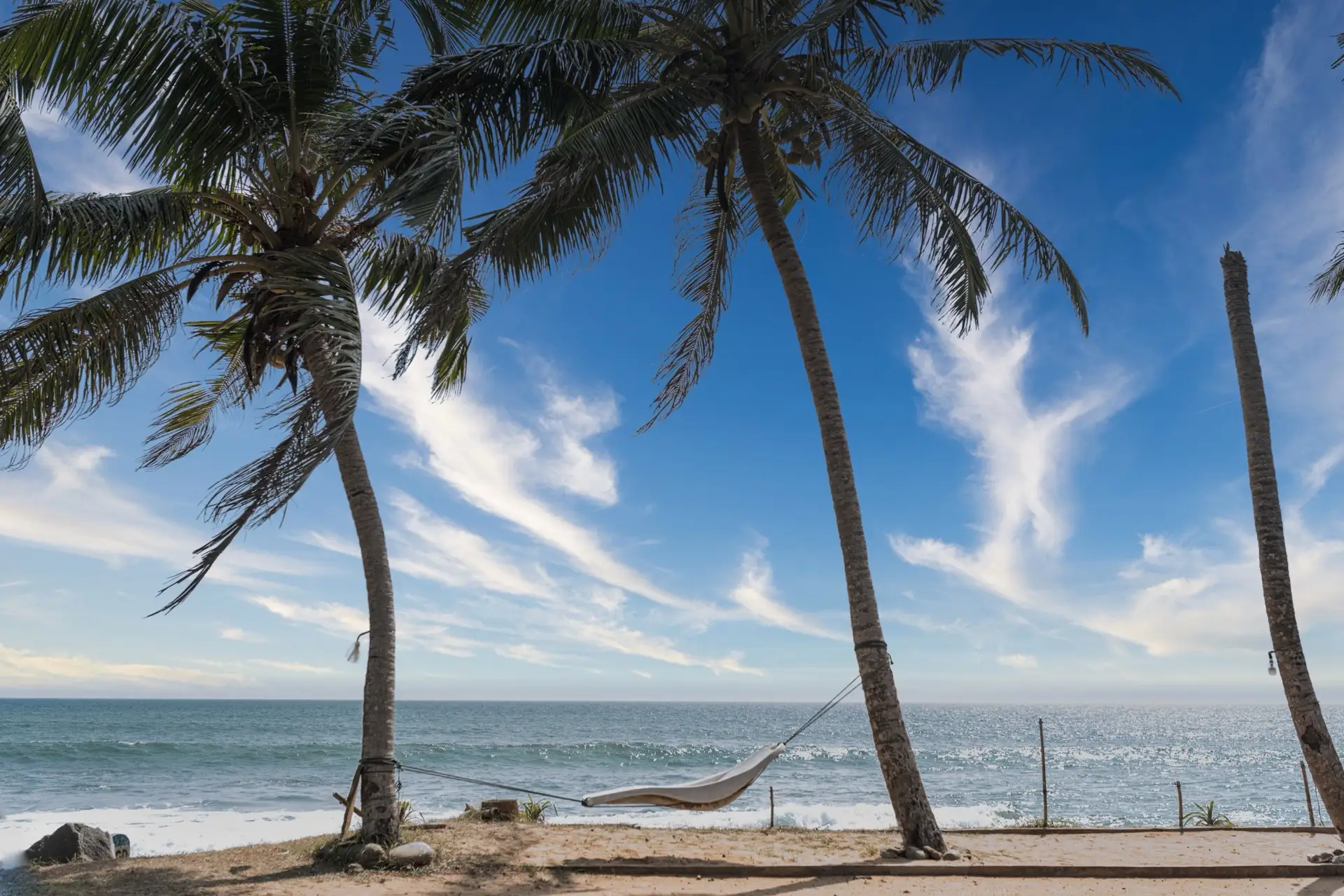 A hammock slung between coconut palms on the sand at Hikkaduwa, with surf breaking behind and a bright sky above.