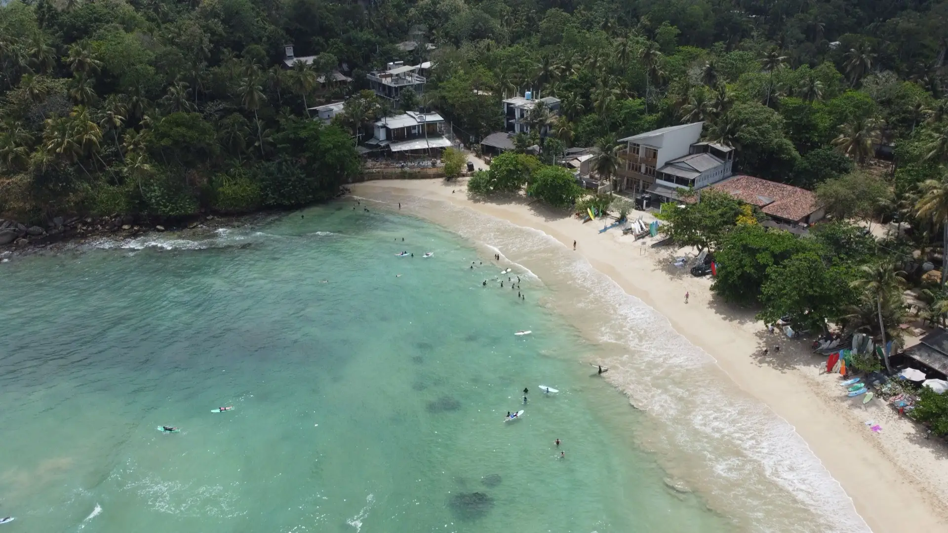 A wide sandy horseshoe bay at Hiriketiya with surfers in the shallows and coconut palms behind the sand.