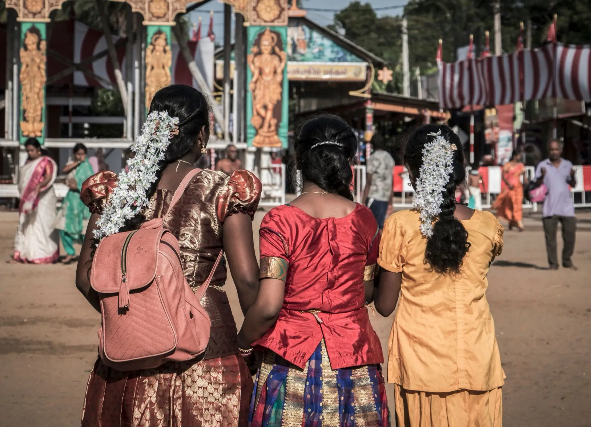 Three women in sarees with jasmine in their hair walking towards a Hindu temple gateway during a festival in Jaffna.
