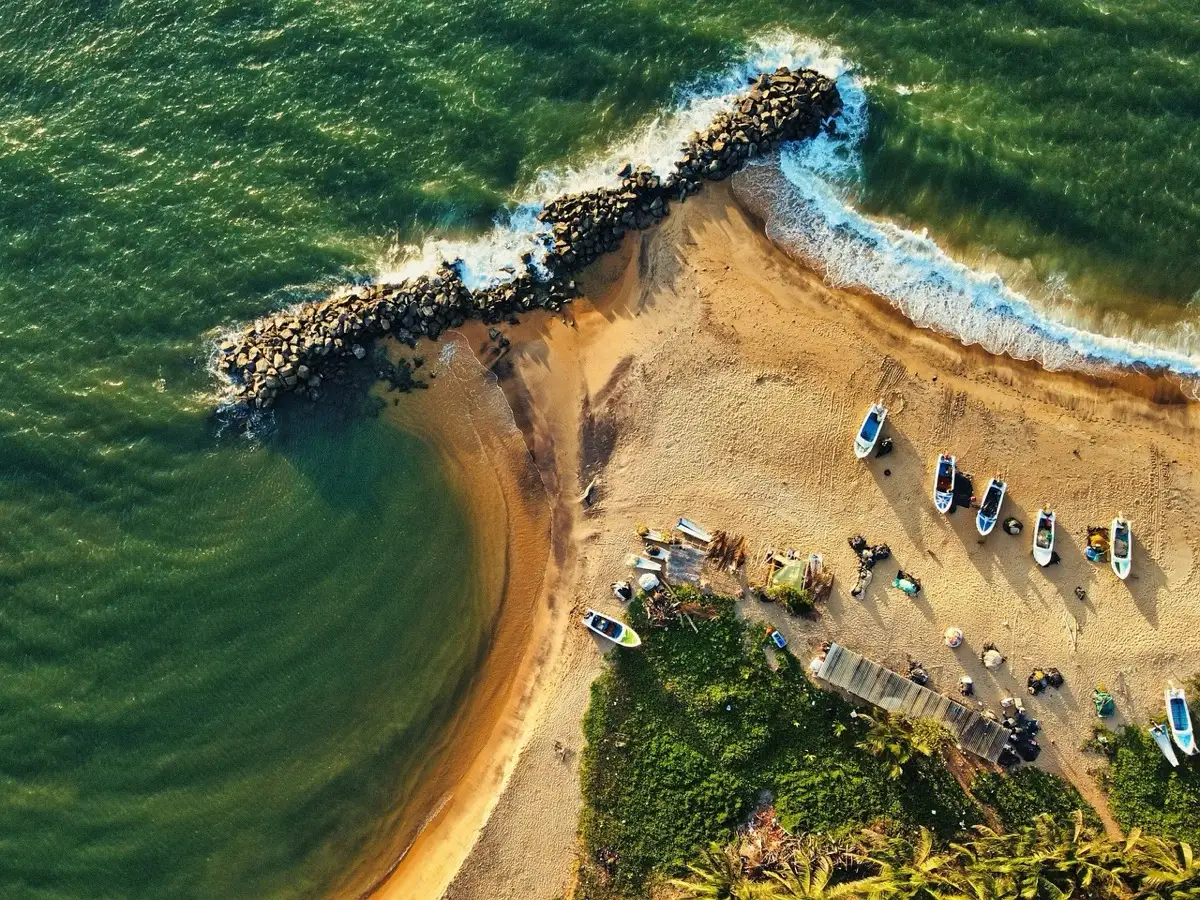 Aerial view of the beach at Kalutara, with a rock groyne running out into green water and fishing boats drawn up on the sand.