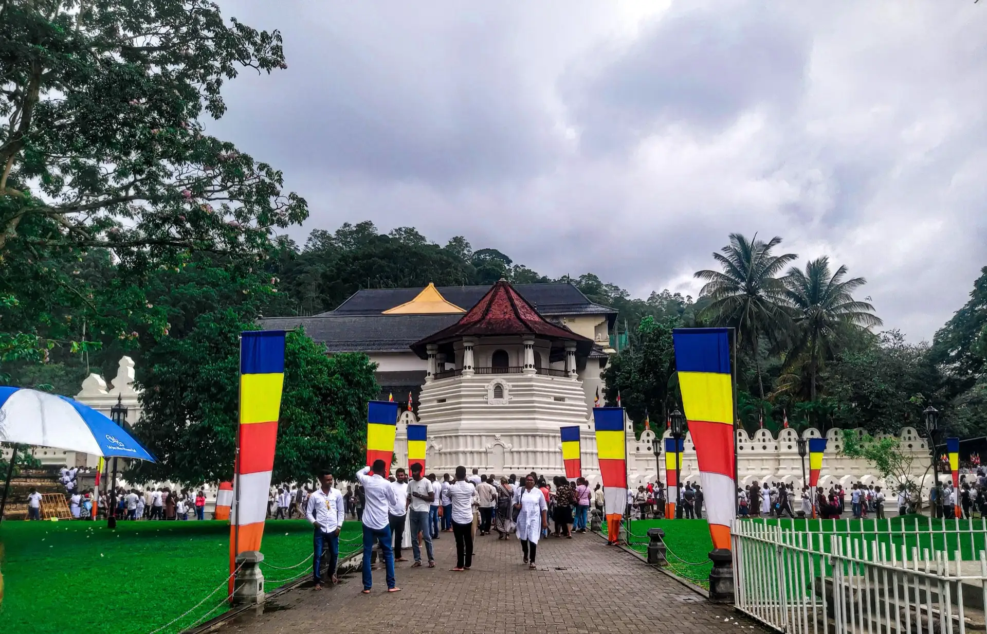 Buddhist flags lining the walkway to the octagonal Pattirippuwa of the Temple of the Tooth in Kandy, with people crossing the forecourt under a grey sky.