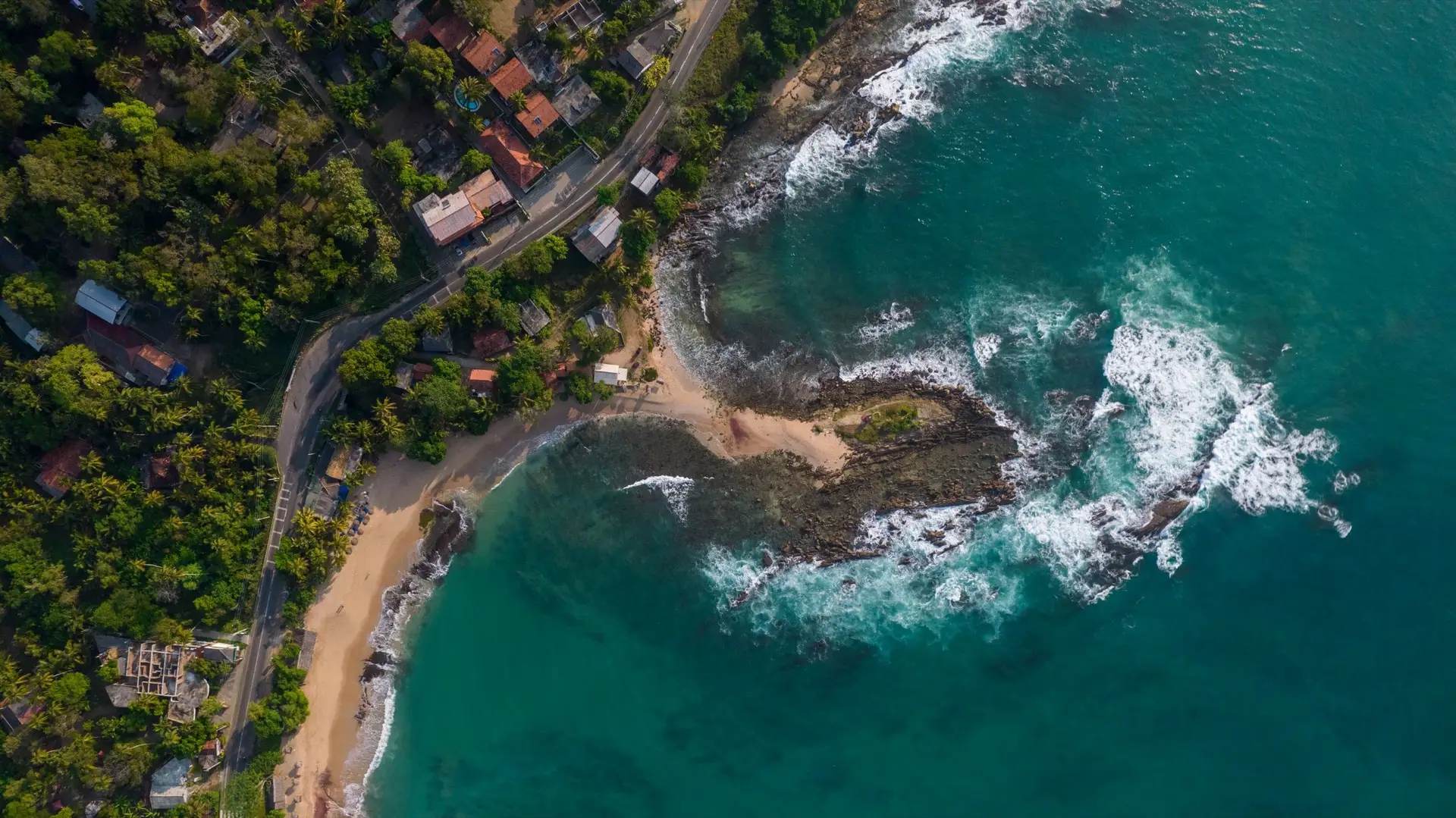 A long curve of sand at Tangalle backed by coconut palms, with surf running up the beach and rocky headlands in the distance.