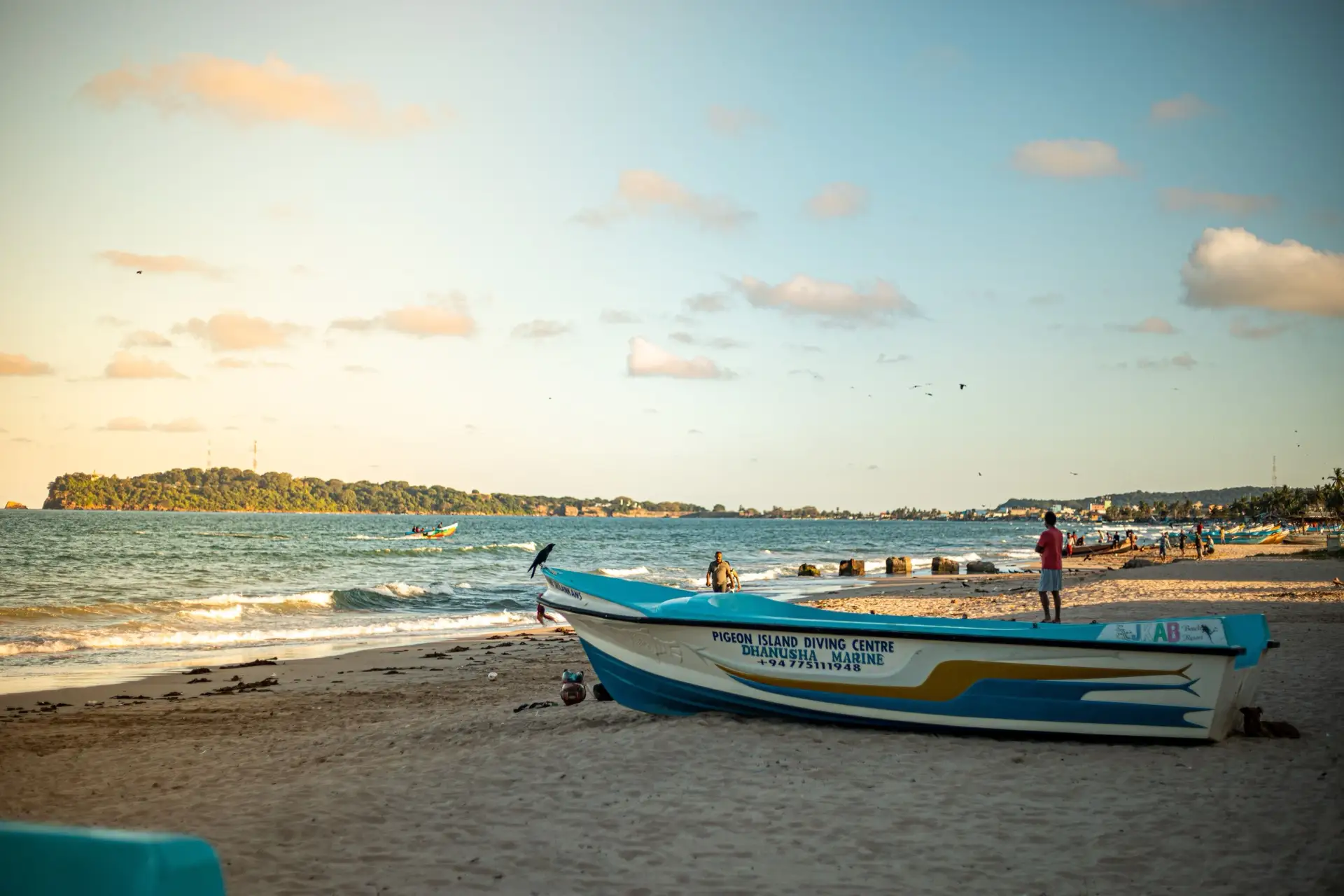 A fishing boat on the sand at Trincomalee in late afternoon light, with a wooded headland across the bay.