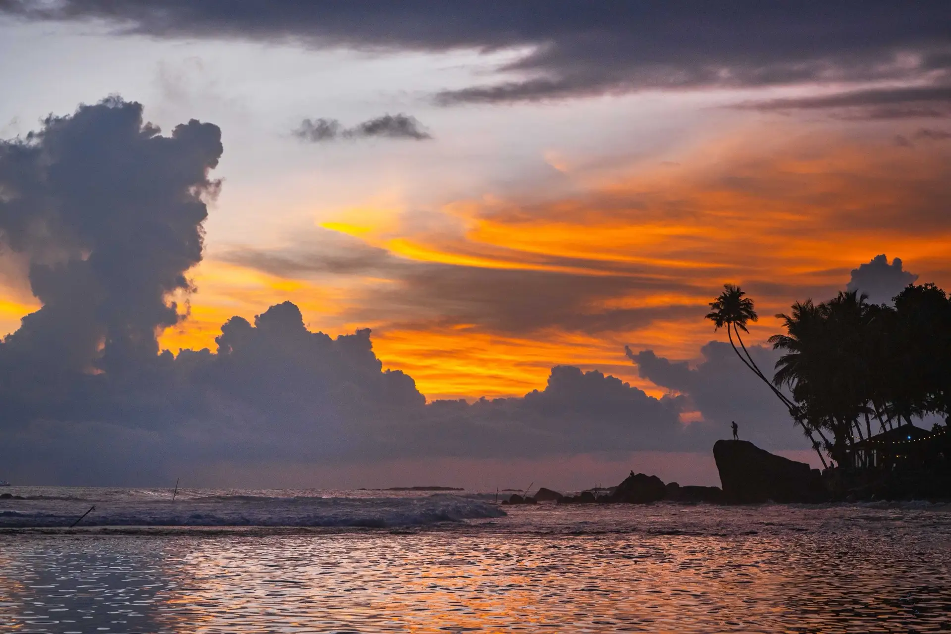 Sunset over the rocks at the end of Unawatuna bay, with a leaning palm silhouetted against orange cloud.