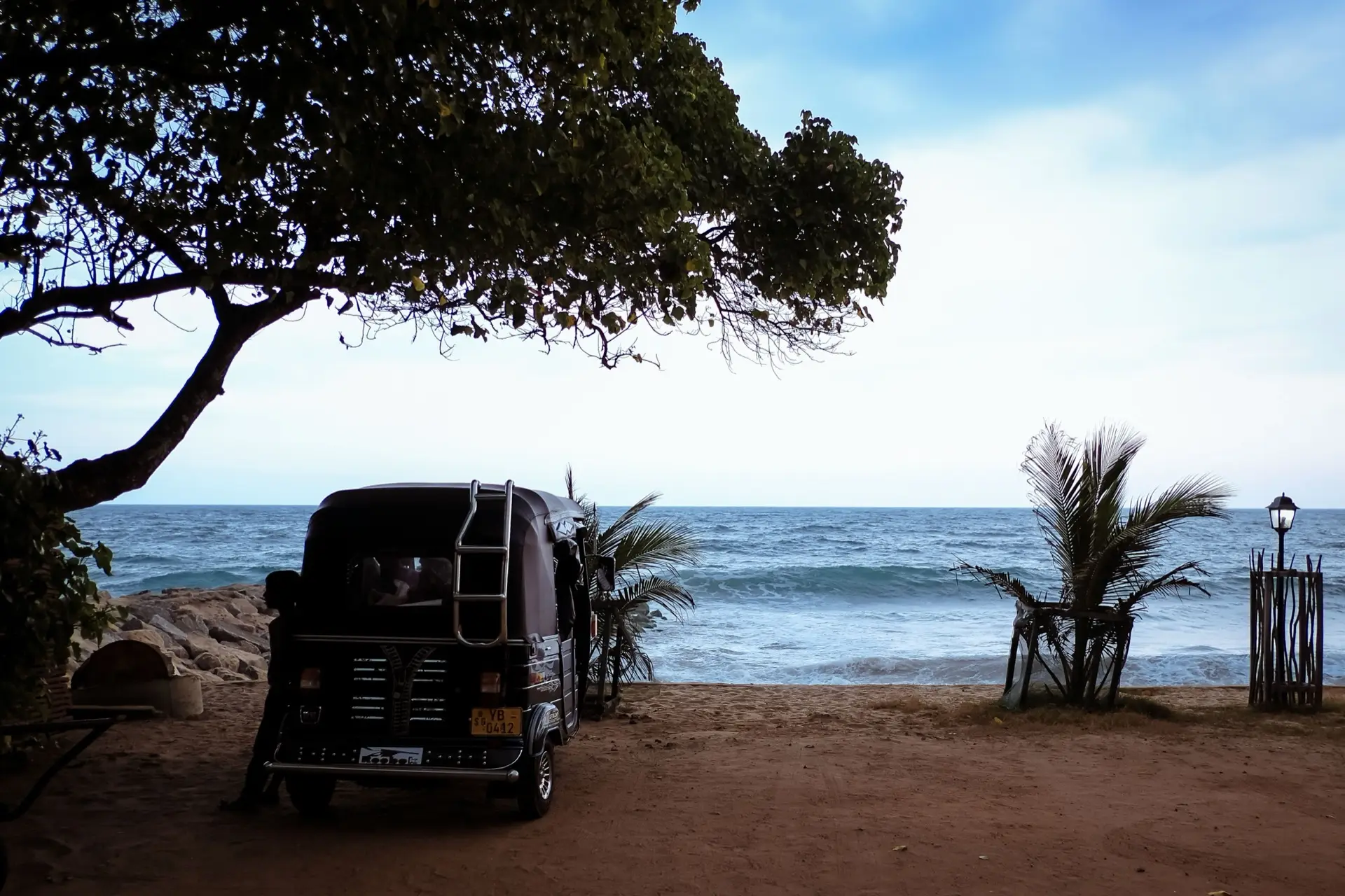 A tuk-tuk parked on sand under a leaning tree, the Indian Ocean behind it.