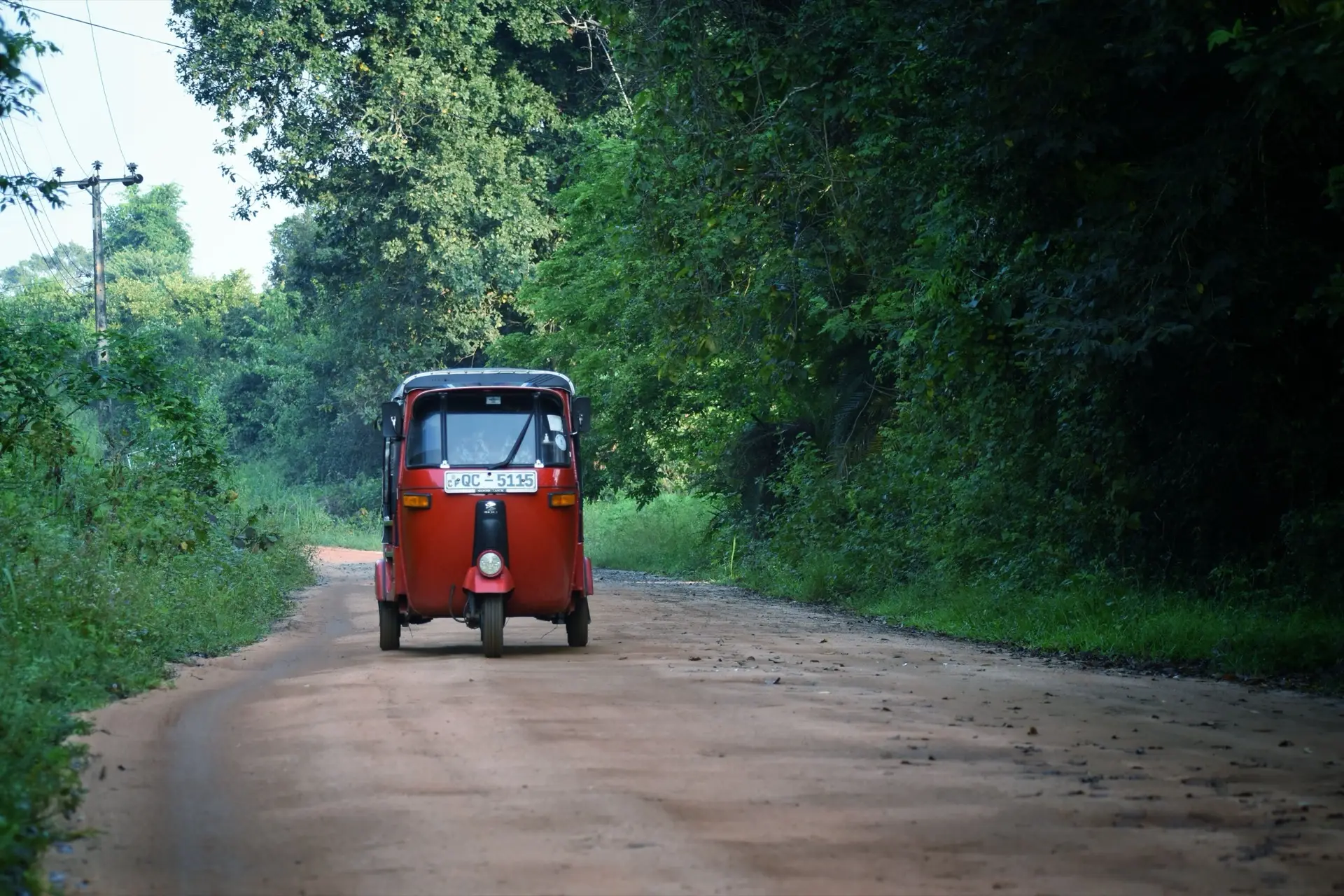 A red tuk-tuk on an empty rural road near Sigiriya, palms and paddy on both sides.
